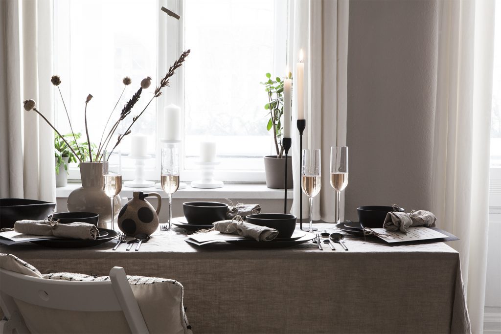 Gedeckter Tisch mit rustikalem, schwarzem Geschirr, Gläser mit Sekt, Leinentischdecke in Natur, Trockenblumen in beiger Steingutvase vor einem Fenster