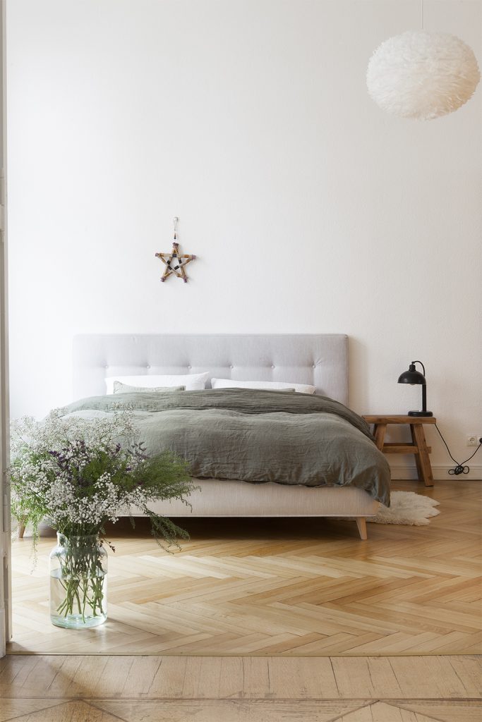 Hellgraues großes Bett mit Stoff bezogen, Holzstern an der Wand, Nachttisch mit Lampe, großer Sommerstrauß in Bodenvase, weiße Feder-Deckenlampe 