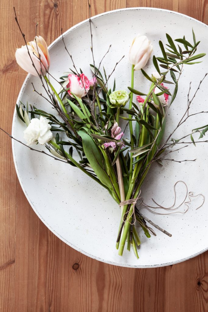 Frühlings Blumenstrauß mit Lederband auf großer heller Schale mit Französischer Tulpe, Papageientulpe, Hyazinthen, Ranunkeln, Birkenzweige, Olivenzweige