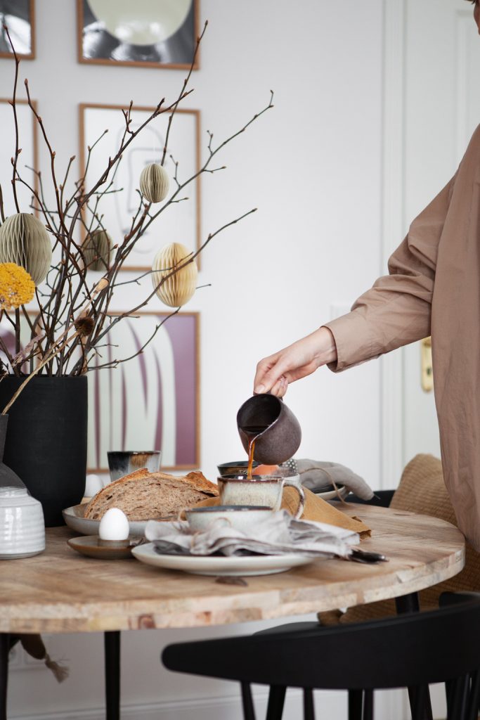 Kaffee in braunem rustikalem Krug wird in Keramikbecher eingegossen, Skandinavisch natürliche Ostertafel an rundem Holztisch mit Brot, Trockenblumen in schwarzer Vase, Bilderwand im Hintergrund