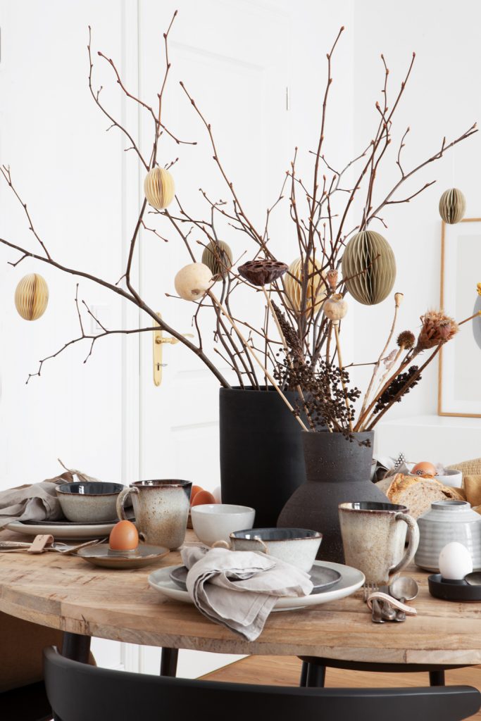 Skandinavisch natürlich gedeckte Ostertafel mit rustikalem Geschirr, Trockenblumen und Geäst in schwarzer und dunkelbrauner Vasen an rundem Holztisch