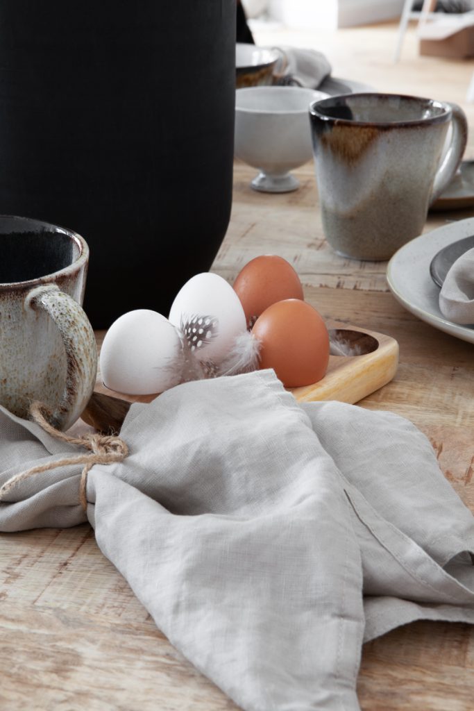 Eierhalter aus Holz mit Eiern, braunem Keramikgeschirr, heller Leinenserviette auf Holztisch