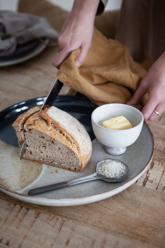 Brot anschneiden auf blau braunem Keramikteller mit Butterschale und Salzlöffel, Hand mit Leinentischtuch schneidet Brot an