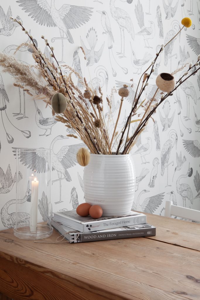 Weiße Keramikvase mit Trockenblumen und Papierostereiern auf Büchern, Glaswindlicht, im Hintergrund Vogel Mustertapete, auf rustikalem Holztisch