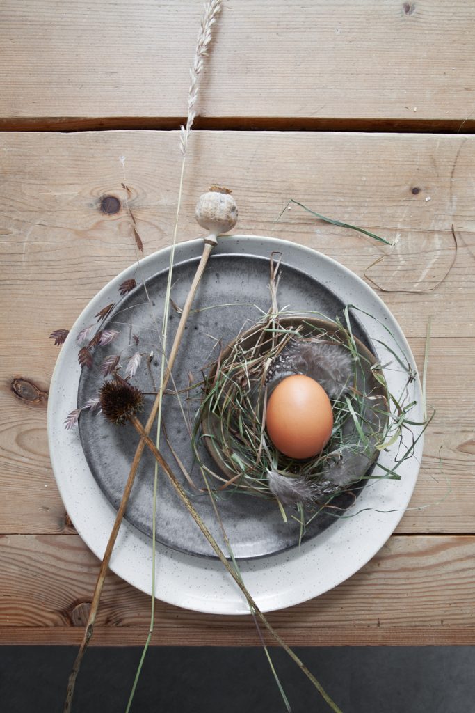 Ei mit Stroh und Feder auf braunem Keramikteller und weißem Keramikteller, Trockenblumen, auf rustikalem Holztisch