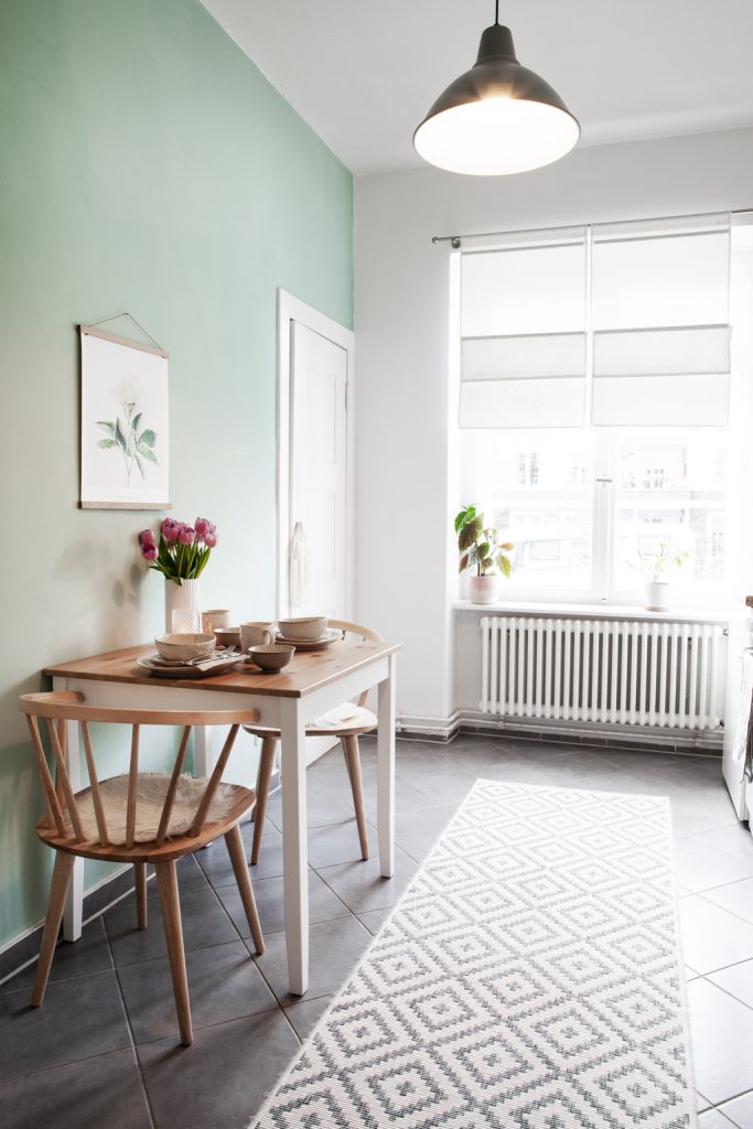 Helle Küche mit mint-farbener Wandfarbe, kleinem Esstisch mit zwei Holzstühlen, einem großen gemusterten Teppich auf grauen Fliesen
