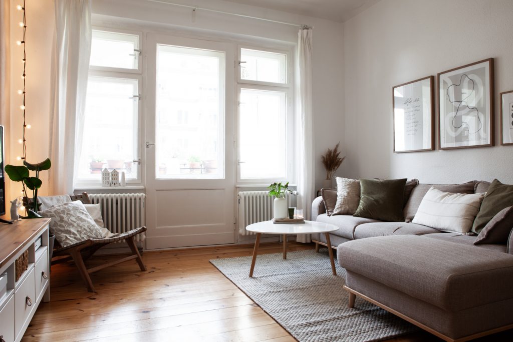 Wohnzimmer mit großem hellem Sofa mit Kissen und Bilder darüber, weißem Couchtisch, flauschigem Teppich, Loungesessel mit Kissen und weißem Sideboard