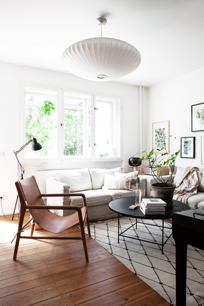 Helles Wohnzimmer mit großer weißer Deckenlampe, braunem Lederstuhl und sandfarbener Couch mit Eichenparkett