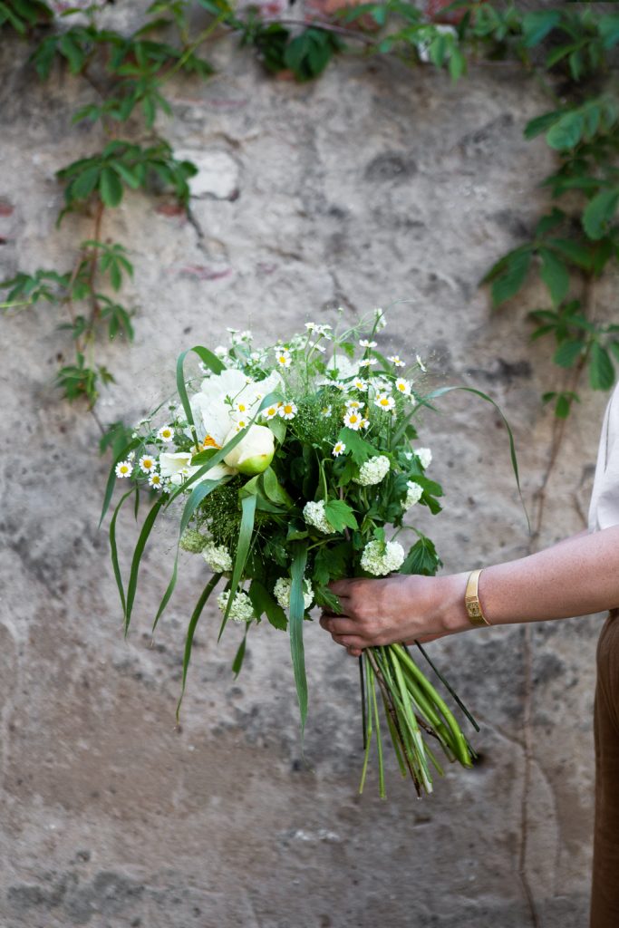 Weißer Sommerstrauß aus Gräsern, Kamille, Pfingstrosen, Schneeball in der Hand gehalten von einer Frau vor alter Mauer mit Efeu berankt
